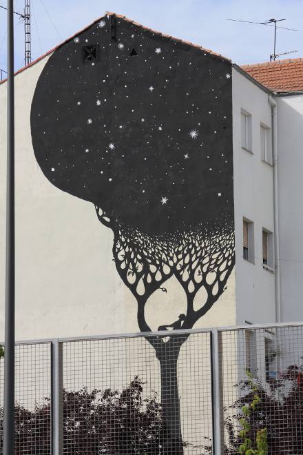 foto di un Murale sulla parete di una casa di un quartiere popolare di Madrid. Al di là dell'inferriata un uomo sdraiato su un ramo guarda i rami dell'albero pieni di stelle